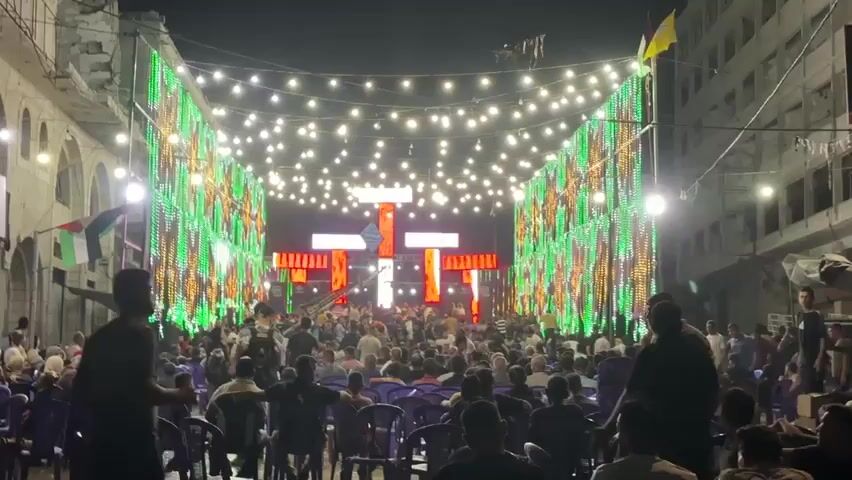 The video opens with a wide shot of a street at night, decorated with numerous strings of lights and vertical light displays on either side. A large crowd of people is gathered in the street, facing a stage at the far end. The stage has a large, abstract, red and white illuminated structure. People are seated on chairs in the foreground, and many more are standing behind them, closer to the stage. The overall atmosphere appears festive, possibly a celebration or an outdoor event. As the video progresses, the lighting displays on the sides of the street change color, cycling through yellow, red, and blue. The stage lighting also shifts in intensity and color. There are flags visible, including what appears to be the Palestinian flag and another yellow flag, hanging from buildings. The buildings themselves seem aged, with visible architectural details. Several individuals are standing in the foreground, some appearing to be security personnel, observing the crowd. The camera remains relatively stationary throughout the clip, focusing on the scene unfolding in the street.