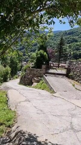The video shows a view of a stone village built into a hillside. The camera pans from left to right, showing a narrow paved road leading uphill towards a closed metal gate. Beyond the gate, a stone wall supports a building. Further to the right, more stone buildings with tile roofs are visible, situated on the hillside. The landscape is lush and green with trees and vegetation covering the surrounding hills. The sky is clear and blue.