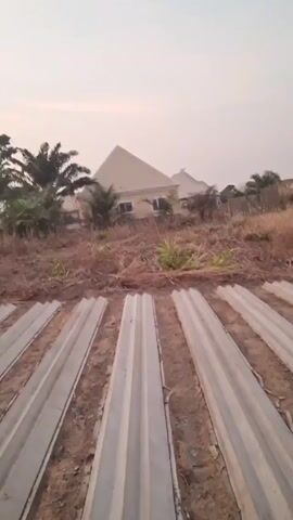 The video shows several long, narrow, gray concrete structures, possibly fence posts or building components, lying horizontally on bare dirt ground. The pieces are ribbed. Behind them is a patch of dry grass and weeds. In the background are houses, suggesting a residential area or construction site. The sky is overcast. There are no people or actions visible in the video.