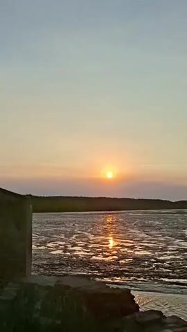 The video pans across a coastal landscape at sunset. A person sits on a wall in the foreground, looking out at the water. The sun is setting over the horizon, casting a warm glow on the scene. In the middle ground, there is a body of water with several boats docked along the shore. In the background, there is a line of trees and vegetation. The overall atmosphere is peaceful and serene.