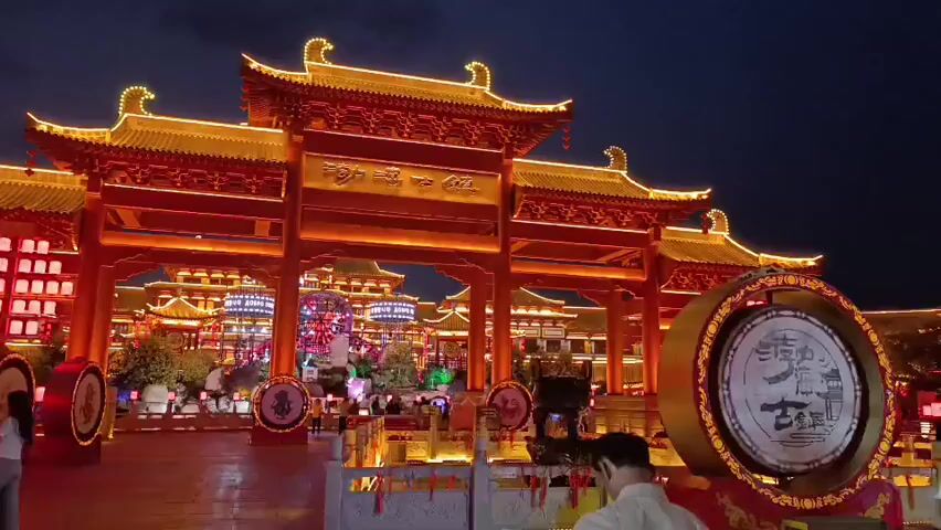 The video captures a scene at night in front of a traditional Chinese architectural structure, possibly a gate or a temple complex, adorned with bright red and gold decorations and illuminated by numerous lights. In the foreground, a large, cylindrical drum with intricate designs is prominently displayed on the right. Two people are visible in the foreground, one standing and facing away from the camera, appearing to be observing something to their right, while another person, a woman with blonde hair, is partially visible on the far right, also looking towards the right. To the left, two individuals, a man in a white shirt and a woman in a light-colored top and dark pants, are walking towards the structure. In the background, the illuminated structure features multiple tiered roofs and ornate detailing. Lanterns and other decorative lights are spread throughout the scene, contributing to a festive atmosphere. Further in the background, other traditional buildings and structures can be seen, also lit up. The sky is dark, indicating it is nighttime. The overall impression is of a vibrant and culturally rich environment, likely a tourist attraction or a location for a celebration.