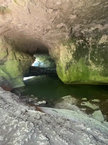 The video shows the interior of a cave or tunnel, with water and rocks on the ground. The walls and ceiling of the cave are rocky and covered in some areas with green moss or algae. The water appears shallow and still, reflecting the cave’s ceiling. Light enters the cave from an opening at the far end. A person can be briefly seen walking in the distance near the cave opening. The camera appears to be held by someone standing inside the cave, panning slightly from side to side to show the features of the cave interior.