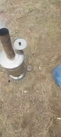 The video begins with a top-down view of a metal stove with smoke emitting from its chimney. The stove is positioned on dry grass and dirt. The camera then pans upwards and to the right, revealing a vast expanse of water, possibly a lake, under an overcast sky. The shoreline is sparsely covered with dry grass. In the final frames, a dark-colored car is visible parked near the water's edge, partially in the frame. The overall scene suggests an outdoor setting, likely a remote or natural location.