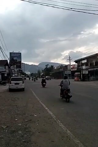 The video shows a medium shot of a street in what appears to be a small town or city during daylight hours. The sky is overcast with grey clouds. A two-lane road runs through the center of the frame, with traffic moving in both directions, including cars and motorcycles. The road is lined with buildings on both sides, which appear to be small businesses or shops. A large billboard featuring an advertisement is visible on the left side of the road.  A mountain range can be seen in the distance. The roadside is somewhat littered with debris and appears unkempt. Electrical lines are strung overhead, crossing the sky. The scene depicts a typical day in a small town with moderate traffic and cloudy weather.