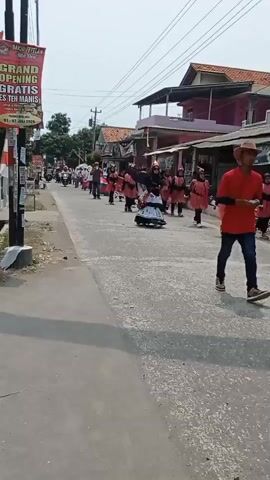 A group of people, some dressed in decorative red and white costumes and masks, are marching down a paved street. The marchers are in single file, and some are holding flags. A motorcycle with two passengers rides past them. Buildings line the street on both sides. A banner advertising a "GRAND OPENING GRATIS ES TEH MANIS" is visible on the left side of the frame. The sky is overcast.