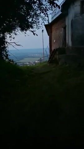 The video is a low-angle, eye-level, moving shot that captures the nighttime view from a hilltop, overlooking a distant town with illuminated lights. The camera pans from left to right, revealing the vast expanse of the town and the surrounding landscape. On the right side of the frame, a building with a balcony and an attached staircase is visible, alongside a tall light pole. Lush greenery and dark trees partially obscure the left side of the frame. The sky above is a dark, clear twilight blue, and the horizon extends into the distance, suggesting a wide valley or plain.
