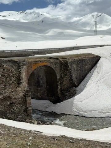 The video shows a low-angle, stationary shot of a stone arch bridge, with a stream flowing underneath it. The surrounding landscape is snowy, with snow covering much of the ground and extending up to the road. In the background, there are snow-capped mountains under a partly cloudy sky. A power line is visible on the mountainside. A few small buildings are seen along the roadside. The camera remains static throughout the video.