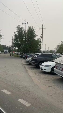 This video captures a drive through an industrial area in Veteran-2, Russia. The scene is dominated by a mix of urban and natural elements. We see a paved road with white lane markings, indicating a structured environment. Large trees, mostly evergreens and deciduous varieties, line the sides of the road, suggesting a location with significant greenery within or adjacent to the industrial zone.

Several vehicles are visible throughout the video. In the initial frames, a row of parked cars is seen, with a white SUV prominently featured as it drives past a guardhouse. Further on, other vehicles are parked and driving through the area, hinting at ongoing activity. The presence of a guardhouse with a barrier suggests an entry or exit point to a facility, possibly a factory or a secure complex. There are also power lines and poles, typical of infrastructure in such locations.

The weather appears overcast, with a hazy or muted sky, suggesting it could be early morning or late afternoon, or simply a cloudy day. The overall atmosphere seems functional and industrial, with no strong emotional cues conveyed.

There is some visible text. On the guardhouse, a blue sign with text is present, but it is not legible. Another sign with the number "5" is also visible. In the distance, a building with a logo on it can be seen, though the logo itself is unclear. The overall impression is of a typical workday in an industrial or logistical area in Russia.