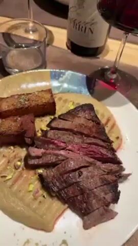 The video shows a table setting in a restaurant, likely during a meal. The table is wooden with individual place settings. Visible on the table are several dishes: one with sliced steak on a bed of sauce, accompanied by fried cubes; another with what appears to be meat and vegetables in a dark, rectangular dish; and a third with fish and vegetables in a light-colored sauce. There are also glasses of red wine and water. People can be seen seated at the table, interacting with the food and drinks. The overall atmosphere suggests a dining experience.