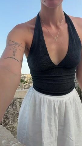 A young woman with blonde hair pulled back and a dark halter top and white skirt stands on a stone ledge overlooking a city of ancient stone buildings. She has a tattoo of a fairy on her left arm and bracelets on her right wrist. The sky is blue and clear. She turns her head and body to face the camera, then turns away, and then faces the camera again.