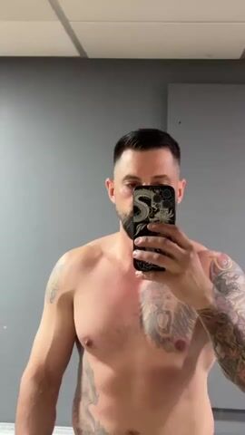 A man is taking a shirtless selfie in front of a gray wall. He has a beard, short dark hair, and tattoos on both arms and his chest. He flexes his right bicep, holding a phone with a black and white patterned case in front of his face. The environment is a plain room with gray walls and a ceiling with fluorescent lights.