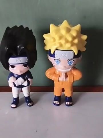 The video shows two plastic figurines of the anime characters Sasuke and Naruto standing on a white surface. Sasuke is on the left, with black hair, a blue and white outfit, and a headband. Naruto is on the right, with spiky yellow hair, an orange jumpsuit, and a headband. They are both facing forward, their arms posed in front of them. The background is a muted green wall with a wooden trim. The camera appears to be still. There is some hammering noise in the background.