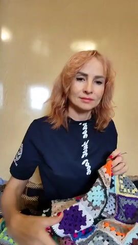 A woman with short, blonde hair is sitting in front of a light-colored wall. She is wearing a dark blue t-shirt and is holding a colorful crocheted blanket made of many square granny squares. She is in the process of folding or displaying the blanket, which features a variety of bright colors like pink, blue, green, orange, and purple, interspersed with neutral colors like gray, beige, and white. Her hands are visible as she manipulates the fabric. To her left, there are some folded items, possibly more fabric or yarn, and a woven basket. The lighting in the room is soft and appears to be coming from the front and slightly above.
