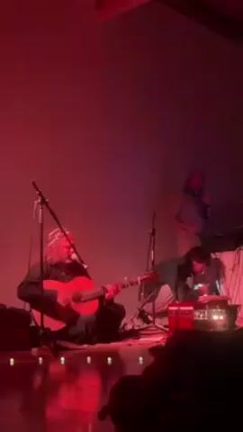 The scene is a dimly lit stage with a red hue, likely a concert setting. On stage, a man with long white hair sits cross-legged on the floor, playing an acoustic guitar. He is positioned in front of a microphone, suggesting he is also singing. To his right, another individual is bending over equipment on the floor, appearing to adjust something. Behind them and to the right, a third man stands at a keyboard or synthesizer, facing forward but angled slightly away from the camera. There are additional microphones and equipment cables visible on the stage. In the foreground, the silhouettes of audience members can be seen. The background is a plain, light-colored wall. A laser light beam briefly flashed across the scene, adding to the concert atmosphere.