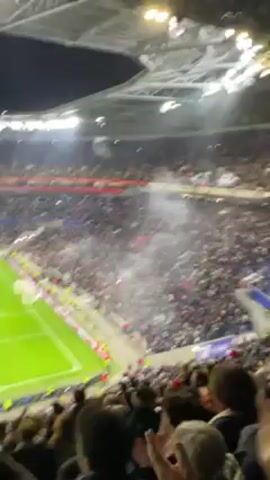 The video shows a large crowd of people at a stadium, likely a soccer game, with smoke or flares visible in the stands. The camera pans across the field and the crowd, capturing the excitement and energy of the event. People are cheering and clapping, their arms raised in the air, and some are recording the scene with their phones. The environment is brightly lit, with the stadium's lights illuminating the field and the audience.