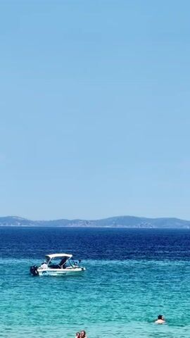 This vertical video showcases a beautiful summer day at a beach in Sithonia, Greece. The scene is dominated by a vast expanse of clear, bright blue sky, transitioning to a deep blue sea in the middle ground, and then to a vibrant turquoise sea closer to the shore.

In the turquoise water, several people are visible enjoying a swim. In the foreground, there are a few individuals, some with their heads above the water, suggesting a relaxed and leisurely atmosphere. Further out, a white motorboat with a canopy is cruising on the deeper blue water. The boat appears to have a few people on board, enjoying a ride. In the far distance, a hazy, mountainous coastline is visible, hinting at the picturesque landscape of Sithonia.

The bright sunlight and clear sky suggest it is a warm and sunny day, likely midday or early afternoon. The overall mood is serene and idyllic, capturing a typical summer vacation scene at a Greek beach. The water is calm, with gentle ripples on the surface.

Visible text on the side of the boat includes the words "SUPER BOAT".