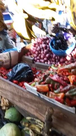 The video shows a market stall in what appears to be an outdoor market. There are bunches of ripe plantains hanging overhead. Below, on the stall itself, are piles of red onions, habanero peppers, tomatoes, and greens in a blue bowl. A person's hand reaches up and touches the plantains. There are other people and boxes visible in the background, indicating a busy market environment.
