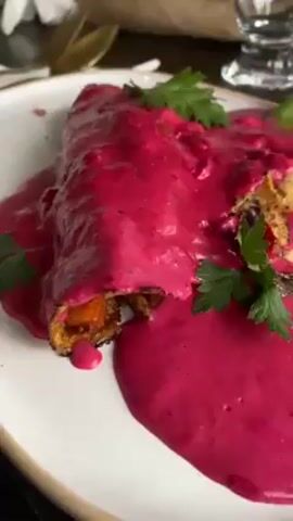 The video showcases two distinct food items served at a restaurant. The first is a rolled dish, possibly a crepe or enchilada, generously covered in a vibrant pink sauce and garnished with fresh parsley. The second dish is presented in a small, dark skillet, filled with a mixture of chopped seafood, likely shrimp or crab, adorned with lemon wedges. The dishes are displayed on a wooden table setting, which also includes beverages, such as a can of 7UP, wine glasses, and a bottle of wine. People are visible in the background, suggesting the food is being served in a social setting.