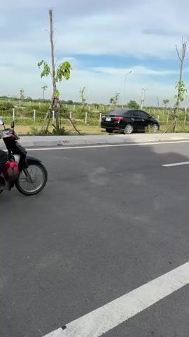 A person is riding a motorcycle down a paved road. The motorcycle is red and the rider is wearing a beige jacket, dark pants, and an orange helmet. To the left of the road, there is a black car parked on the side, and further back, there are some trees and power lines. To the right of the road, there is a sidewalk with some parked motorcycles and a green canopy. The sky is blue with some clouds.
