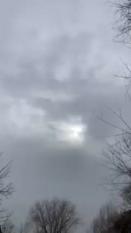 The video begins with a view of a cloudy sky, the sun faintly visible through the clouds. The camera then pans down to reveal a rural landscape, showing a dirt path, leafless trees, and a small, simple building. The scene appears to be in a village or rural area. There is some snow on the ground. A finger briefly covers the lens before the view clears again. There is a pile of brush next to the path. A donkey is visible, grazing in the distance. The overall tone of the video is muted and somewhat overcast due to the cloudy weather.