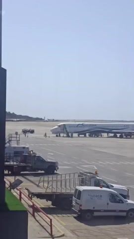 The video shows an airplane parked on a tarmac. The plane is white with a dark blue tail and a dark blue stripe along the length of the fuselage. The name "Venezolana" is written on the side of the plane. Stairs are connected to the front of the plane and several people are standing around the base of the stairs. The sky is clear and blue. In the foreground, there are several vehicles, including a white van with the word "Butaca" on the side. The camera zooms out slightly to reveal that the scene is being viewed from inside a building through a window.
