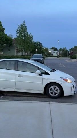The video shows a white Toyota Prius parked in a parking lot. A man wearing sunglasses and a black cap is sitting in the driver's seat. He looks toward the camera and briefly smiles. The sky is dusky, and there are trees and buildings in the background. Another dark-colored car is parked behind the Prius. Streetlights are visible in the background.
