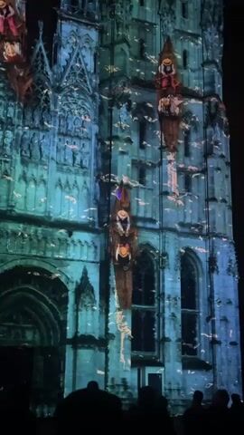 A video shows a nighttime light projection onto the facade of a large, Gothic-style stone building, likely a cathedral or church. The projection covers the entire visible front of the building, transforming it into a vibrant, animated scene. The building's architectural details, such as windows, arches, and intricate carvings, are visible beneath the projected light.

The dominant colors of the projection are shades of blue and teal, giving the building an aquatic or dreamlike appearance. Numerous small, light-colored particles, resembling bubbles or sparkles, are projected across the surface, creating a dynamic, shimmering effect.

Prominently featured in the projection are several wooden boats, each containing one or more figures. These boats appear to be floating or sailing upwards, vertically along the building's facade. Two main boats are clearly visible on the right side of the frame, moving towards the top. Another boat is partially visible on the left side.

The figures within the boats appear to be stylized or animated characters, dressed in various colorful garments, possibly representing historical or mythical personae. Their postures suggest they are seated or standing within the boats.

At the bottom of the frame, the dark silhouettes of several people can be seen, presumably an audience watching the light show. They are indistinguishable but provide context for the scale of the building and the event. The overall scene is dark, with the illuminated building standing out against the night sky.