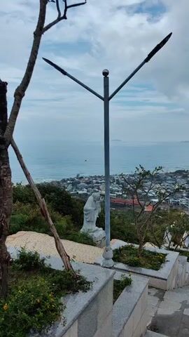 The video captures a scenic overview of a coastal town from a high vantage point, presumably from a hillside or temple complex. The camera pans upwards along a stone staircase lined with statues, with lush greenery and planters flanking the steps. In the distance, the town sprawls along the coastline, with a sandy beach and the ocean stretching out to the horizon under an overcast sky. Wind turbines are visible on the distant horizon, suggesting an environmentally-conscious energy infrastructure. The scene is calm, with a few people walking along the stairs.