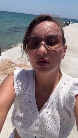 This is a selfie-style video of a young woman standing on a concrete pathway next to a body of water, likely the sea. She is wearing a white crocheted crop top with ruffled sleeves and matching white shorts. She also wears large, tinted sunglasses. The background shows the clear blue sea with gentle waves and the sky. The sun appears to be bright, suggesting a sunny day. The woman smiles and speaks to the camera. There is some sand and debris on the pathway. The location appears to be a coastal area, possibly a beach or a promenade.