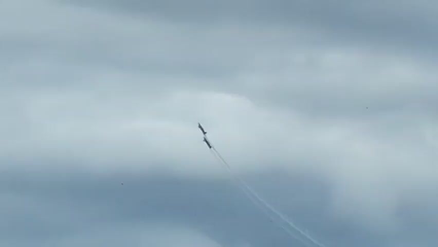 Two dark-colored aircraft fly in formation against a cloudy sky. The lead aircraft is slightly ahead and to the left of the trailing aircraft. Both aircraft are leaving faint trails of smoke, one red and one blue, as they ascend. The aircraft are small in the frame and appear to be performing an aerial maneuver. The sky is filled with grey and white clouds, obscuring any view of the ground. The overall lighting is dim, consistent with an overcast day.