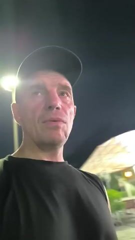 The video shows a bald, Caucasian man wearing a black t-shirt and a black baseball cap. He is speaking directly to the camera, and the background appears to be a street scene at night. There are illuminated signs in Chinese characters, and some blurry figures of people in the background. The man states that his trip to China has ended and he is returning home.