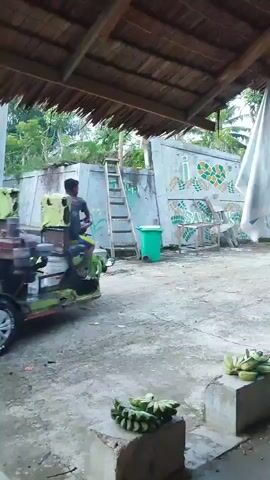 A group of seven men, dressed in casual clothing, are seen pushing a green and white motorized vehicle from the left side of the frame towards the right. The vehicle has a visible engine and a driver sitting on top. The men are exerting force on the back of the vehicle, working together to move it. The environment appears to be an outdoor area with a thatched roof overhead and a concrete ground surface. In the background, there is a building with a mural and some green bins. Bunches of green bananas are placed on concrete blocks in the foreground. The scene suggests a collaborative effort to move the vehicle, possibly due to it being immobile or needing to be positioned.