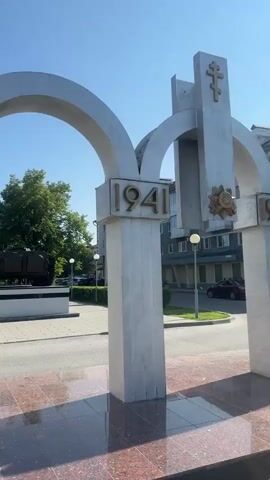 The video shows an outdoor scene on a sunny day. In the foreground, there is a large silver archway and a column with the numbers "1941" displayed. The column has a textured pattern. To the left of the archway, through the opening, a military tank is visible, mounted on a pedestal. The tank is dark green or brown. The ground in front of the archway appears to be paved with red and black marbled stones. In the background, there is a street with cars, buildings, trees, and lampposts. The sky is clear and blue. The camera is positioned at a medium distance from the archway and tank.