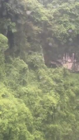 The video shows a lush, green mountainside, possibly filmed from a moving vehicle or elevated position. There is dense vegetation covering the slopes. A small, rocky river can be seen at the bottom of the frame, and the background is slightly obscured by rain or mist. The overall tone is natural and slightly hazy.