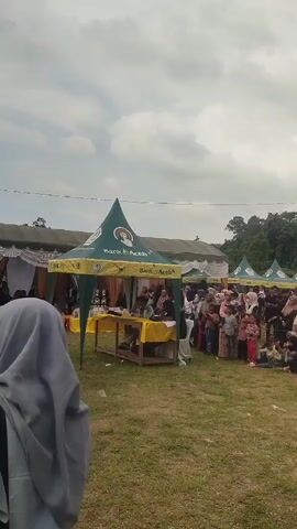 The video shows an outdoor event with several tents set up. In the foreground, a person wearing a grey hijab is visible from the back. The main focus is on a green tent with "Bank Aceh" branding. Underneath this tent, there is a wooden table covered with a yellow cloth. Several people are seated at the table, appearing to be participants or judges. One young man in a light green shirt is leaning forward, seemingly speaking or observing something. Behind him, another person is writing. In the background, a crowd of people, including many women and children dressed in traditional attire, are gathered, watching the proceedings. To the left of the main tent, there are some trophies displayed on a table. The ground is covered with grass. The sky is overcast.