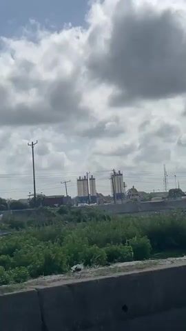 The video is taken from the perspective of someone riding in a truck on a highway. The highway has a concrete barrier in the median. The truck passes a piece of heavy machinery, a crane, and a cement mixing facility in the background. The weather is partly cloudy. The environment appears industrial with grassy areas. There is nothing to indicate anything extraordinary or unusual is happening. The video captures a drive-by view of an industrial landscape from a truck.