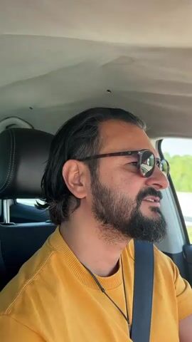 The video shows the point of view of a person driving a car on a highway. The driver is a man with dark hair and a beard, wearing glasses and a yellow sweater. The car interior is visible, including the dashboard, steering wheel, and seats. The exterior view shows the highway stretching into the distance under a clear blue sky. Lush green trees and bushes line the sides of the road. Other cars and trucks can be seen driving on the highway. The sun is shining brightly. The camera appears to be mounted inside the car, capturing the driver's perspective and the scenery passing by. The video also contains music.