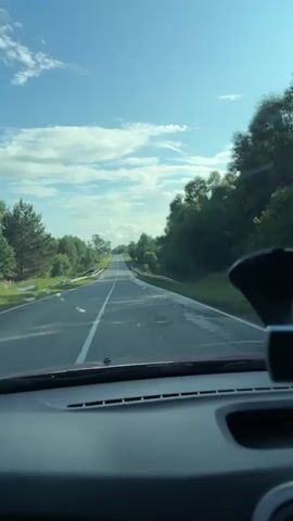 The video is a first-person perspective shot from inside a car. The car is driving on a two-lane asphalt road, with a white line dividing the lanes. The road is surrounded by green trees and vegetation on both sides. The sky is blue with scattered white clouds. There are guardrails along the sides of the road. The dashboard of the car is visible in the foreground, with a dark material and some visible vents. A black object, possibly a dashcam or GPS device, is mounted on the dashboard in the right side of the frame. The sun appears to be shining, casting shadows. The road ahead is straight and appears to curve slightly towards the right in the distance. The scene suggests a drive through a rural or natural landscape.