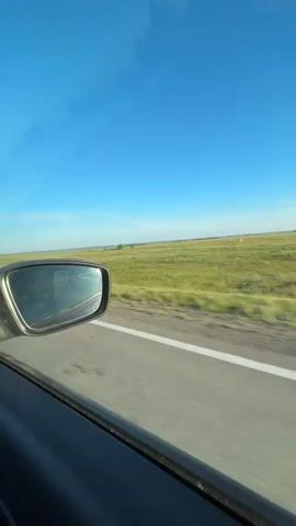 The video is filmed from inside a moving vehicle, looking out the passenger window. The camera is angled slightly upwards, capturing a bright blue sky with a few wispy clouds. In the foreground, the black frame of the side mirror is visible, reflecting the sky and the interior of the car. The road is visible as a grey asphalt strip with a white line along the edge, indicating the direction of travel. To the right of the road, a vast expanse of green and yellow fields stretches into the distance. There are scattered trees and bushes along the roadside and in the fields, and some hints of distant structures or buildings. The impression is of a scenic drive through a rural, possibly agricultural landscape on a clear day. The motion blur indicates the vehicle is in transit.