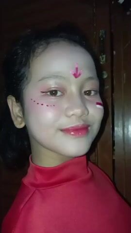 A young girl with dark hair styled in a low ponytail is wearing red clothing and makeup. Her face is painted white, with pink blush on her cheeks, pink eyeshadow, and bright pink lipstick. There is a pink arrow design on her forehead and pink dots below her eyes. A small red and white stripe is painted on her right cheek, representing the Indonesian flag. She is smiling and looking towards the camera. The background appears to be indoors with a wooden door to the right.