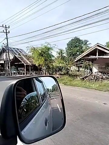 A rearview mirror of a moving vehicle reflects a road, buildings with thatched roofs, trees, utility poles with wires, and a clear sky. The perspective suggests a journey through a rural or semi-rural area. The buildings appear to be small shops or homes with open fronts. A person's arm is visible in the reflection of the car's interior. The video pans slightly to the right, showing more of the environment. A white car and a motorcycle are seen in the distance on the road.