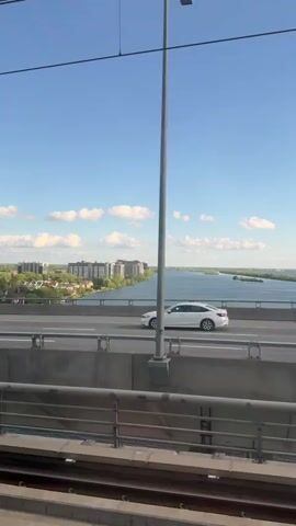 The video shows a white semi-truck driving across a bridge on a sunny day. A commuter train passes in the opposite direction, with passengers visible through its windows. The surrounding environment includes a body of water, urban buildings, and a blue sky with scattered clouds. Overhead power lines are visible throughout the video.