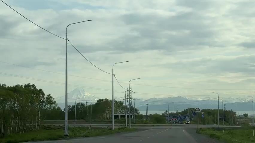 The video shows a road with street lights and power lines. In the distance, there are mountains, some of which appear to be volcanoes. There are a few cars on the road, and a speed limit sign that indicates 40. A round-about sign is also visible. The sky is cloudy. The overall impression is of a somewhat remote, possibly rural or semi-urban area with a striking natural landscape in the background.