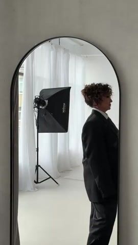A woman with curly brown hair is reflected in a large arched mirror. She is wearing a dark pinstripe suit jacket, a white shirt, and a striped tie. Her head is tilted down, and she is looking slightly to her right. Behind her, reflected in the mirror, is a white room with sheer white curtains and a photography light with a softbox on a stand. To the left of the light, a building with windows is visible through the curtains. The mirror frame is black.