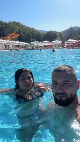 A man and a woman are in a swimming pool. The woman is wearing a bikini and the man is shirtless. They are both submerged in the water, with the man holding a camera to film them. In the background, there are lounge chairs and umbrellas by the pool, and trees and a water slide are visible further in the distance. The water appears clear and blue.
