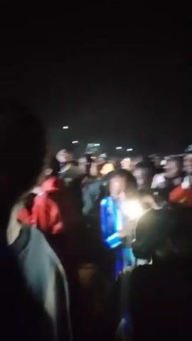 The video shows a crowd of people at an outdoor event, likely a concert or festival, at night. The crowd is diverse in appearance and attire, with many wearing hats, caps, and various colors of clothing. The lighting is poor, with light sources seemingly coming from distant points and some handheld devices. The camera moves through the crowd, capturing glimpses of individuals and their interactions. The music is audible, suggesting the presence of a performing act. The scene is somewhat chaotic due to the movement and density of the crowd.