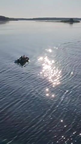 A dark-colored inflatable boat with two people in it moves left to right across a wide body of water, leaving a wake behind it. The sun glares off the water to the right of the boat. Distant land is visible in the background under a light blue sky.