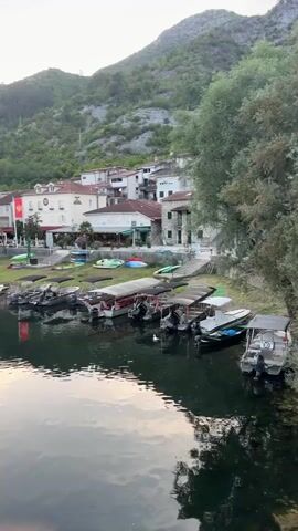The video pans across a waterfront scene at dusk. The foreground shows a body of water with reflections of the sky and surrounding landscape. Along the shoreline, a row of small boats with canopies is docked. Behind the boats, a village with white and light-colored buildings, some with red roofs, is situated at the base of a lush, green, tree-covered mountain. The sky is a soft gray and orange, indicating sunset. The overall impression is a peaceful, serene, and scenic view of a lakeside village.