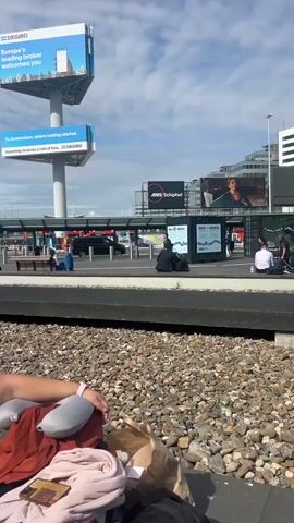 The video shows an adult man in a pilot's uniform walking with a suitcase on wheels through what appears to be a transportation hub, possibly an airport. He walks past two people sitting on a bench. The setting includes buildings with large signs, and a vehicle pulls into the loading area. The ground is covered in small stones and asphalt.