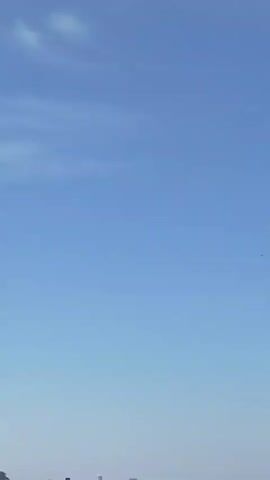 The video shows a clear blue sky with a few wispy white clouds. In the distance, a faint line of buildings is visible at the bottom of the frame, suggesting an urban or suburban landscape. There is a tiny dark object, possibly a bird or a drone, flying in the sky. The camera appears to be held steady, and the focus is on the expansive sky.